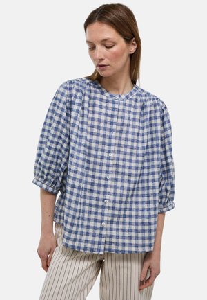 Femme portant un chemisier boutonné à carreaux bleu et blanc avec des manches bouffantes 3/4 et un pantalon beige à fines rayures verticales.