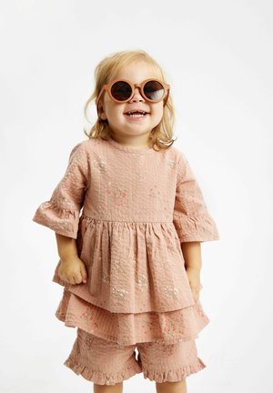 Rosa mönstrad klänning med volangärmar och lager på lager-kjol, matchad med shorts. Barnet bär runda solglasögon och ler.