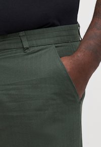 Dunkelgrüne Hose mit Fischgrätstruktur, ausgestattet mit Seitentaschen und einem Bund. Das Material wirkt glatt und strapazierfähig.