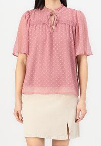 Blouse rose transparente avec texture à pois, décolleté en trou de clé, et manches bouffantes, associée à une jupe en velours côtelé beige clair avec une fente sur le côté.