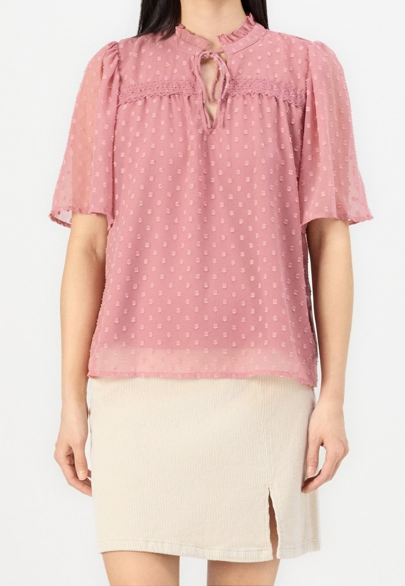 Blouse rose transparente avec texture à pois, décolleté en trou de clé, et manches bouffantes, associée à une jupe en velours côtelé beige clair avec une fente sur le côté.