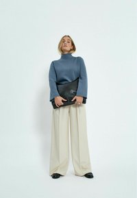 Zils augsta kakla džemperis ar garām piedurknēm; krēmkrāsas platajām biksēm; melna ādas sajūga soma; gaiša fona.