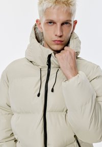 Beige Puffer-Jacke mit Kapuze, ausgestattet mit einem schwarzen Reißverschluss und Zugbandsakzenten. Glatte, gepolsterte Textur mit einem leichten Design.