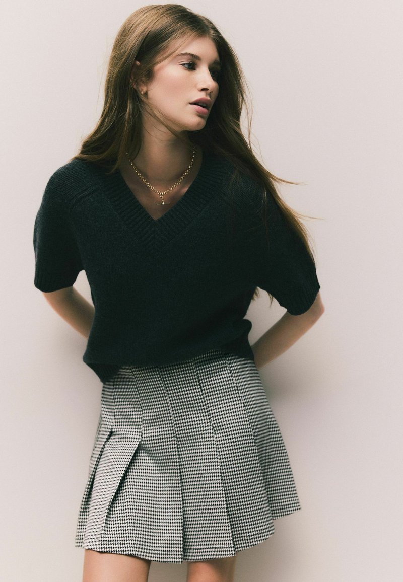 Sort kortærmet sweater med V-hals, parret med en plisseret sort-hvid houndstooth nederdel, der har tekstureret materiale og rene linjer.