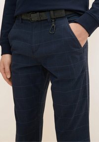 Kalhoty s námořnickým vzorem, které mají jemný kostkovaný design, vyrobené z lehkého materiálu. Součástí je černý opasek a bezpečná kapsa.