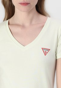 Jasnożółta koszulka z krótkim rękawem i dekoltem w serek, z małą czerwoną, trójkątną łatą z logo "GUESS" na lewej piersi. Miękka tekstura materiału.