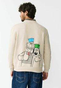 Beiger Pullover mit gerippten Bündchen an Ärmeln und Saum, mit einer Cartoon-Grafik von zwei Charakteren mit bunten Kronen auf der Rückseite.