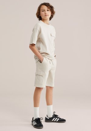 Persona joven de pie con las manos en los bolsillos, vistiendo pantalones cortos beige, camiseta a juego, calcetines blancos y zapatillas Adidas negras con rayas blancas.