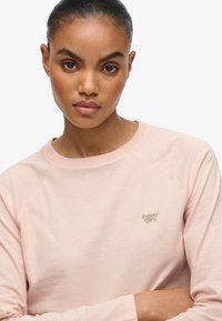 Helles rosa Sweatshirt mit langen Raglanärmeln und Rundhalsausschnitt; verfügt über ein kleines, beiges "Superdry"-Logo auf der Brust. Glatte Stoffoberfläche.