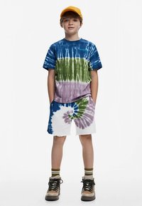 Tricou tie-dye în albastru, verde și mov cu croială lejeră; asortat cu pantaloni scurți albi cu un model colorat în spirale. Cizme maro și șapcă galbenă.