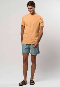 Férfi barack színű rövid ujjú pólót, kék és fehér csíkos rövidnadrágot, valamint sötét szandált visel, egy sima világos háttér előtt áll.