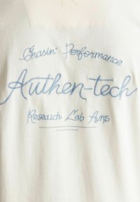 Tessuto bianco con testo ricamato blu: "Chasin’ Performance Authen-tech Research Lab Ams" e un testo più piccolo sottostante che dettagli le aree di innovazione.