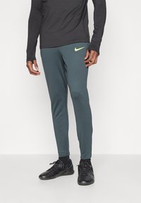 Muške Nike atletske hlače u tamnozelenoj boji, sa uskim krojem, elastičnim pojasom i žutim logotipom na lijevom butu.