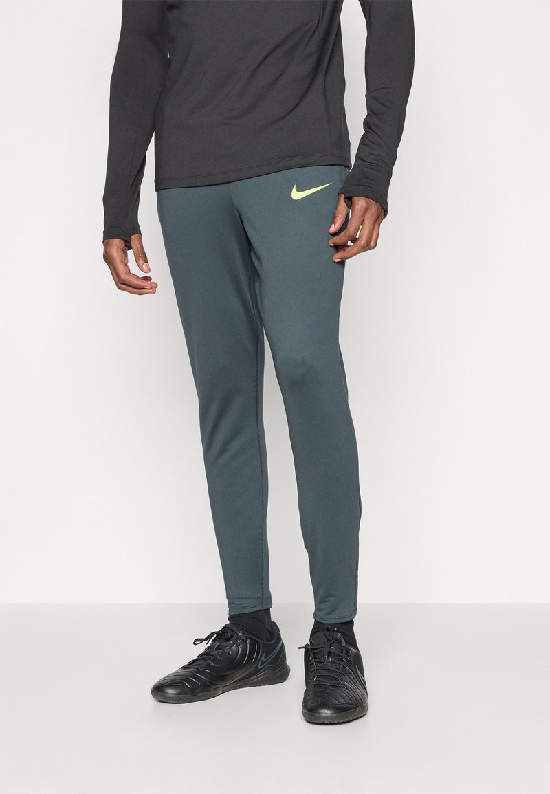 Muške Nike atletske hlače u tamnozelenoj boji, sa uskim krojem, elastičnim pojasom i žutim logotipom na lijevom butu.