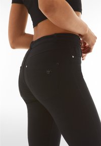 Mustat farkut, joissa on istuva malli, korkea vyötärö ja kaksi takataskua, joissa on pieni logotehoste. Kangas näyttää sileältä ja joustavalta.