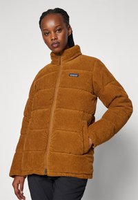 Giubbotto puffer in corduroy marrone con colletto alto, chiusura a zip e tasche laterali. Presenta un design trapuntato e un piccolo logo sul petto.