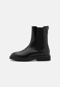 Botas de cuero negras hasta el tobillo con paneles laterales elásticos, punta redondeada y suela de goma texturizada. El diseño incluye una pestaña en la parte posterior.