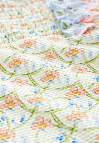 Cvetlična odeja z rožnatimi in modrimi cvetovi na svetlo zeleni teksturirani tkanini, z nabrano obrobo v mehki modri barvi.