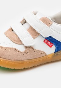 Baskets blanches avec des accents en daim beige et bleu, straps en velcro, boîte à orteils perforée et semelle en caoutchouc transparent. Présente une étiquette de logo rouge.