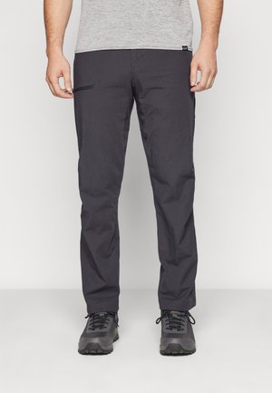 Moški, oblečen v temnosive hlače z ravnimi hlačnicami, z bočno zadrgo na žepu in črnimi pohodniškimi čevlji, stoji na svetlem tlaku.