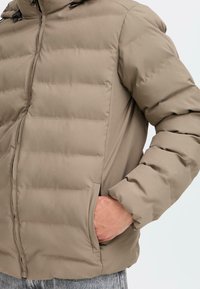 Puffjakke i lys brun, med quiltet mønster, sidelommer og lynlås. Materialet ser blødt og isoleret ud.