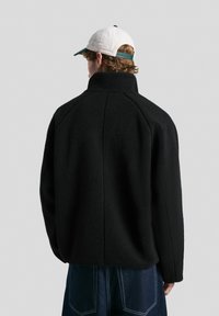 Jachetă din fleece neagră cu guler înalt, cu umeri lăsați și o croială lejeră, asortată cu pantaloni din denim albastri.