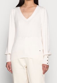 Pull à col en V en maille crème douce avec manches bouffantes. Comprend un ourlet côtelé et des poignets avec des accents de boutons. Associé à un pantalon de couleur claire.