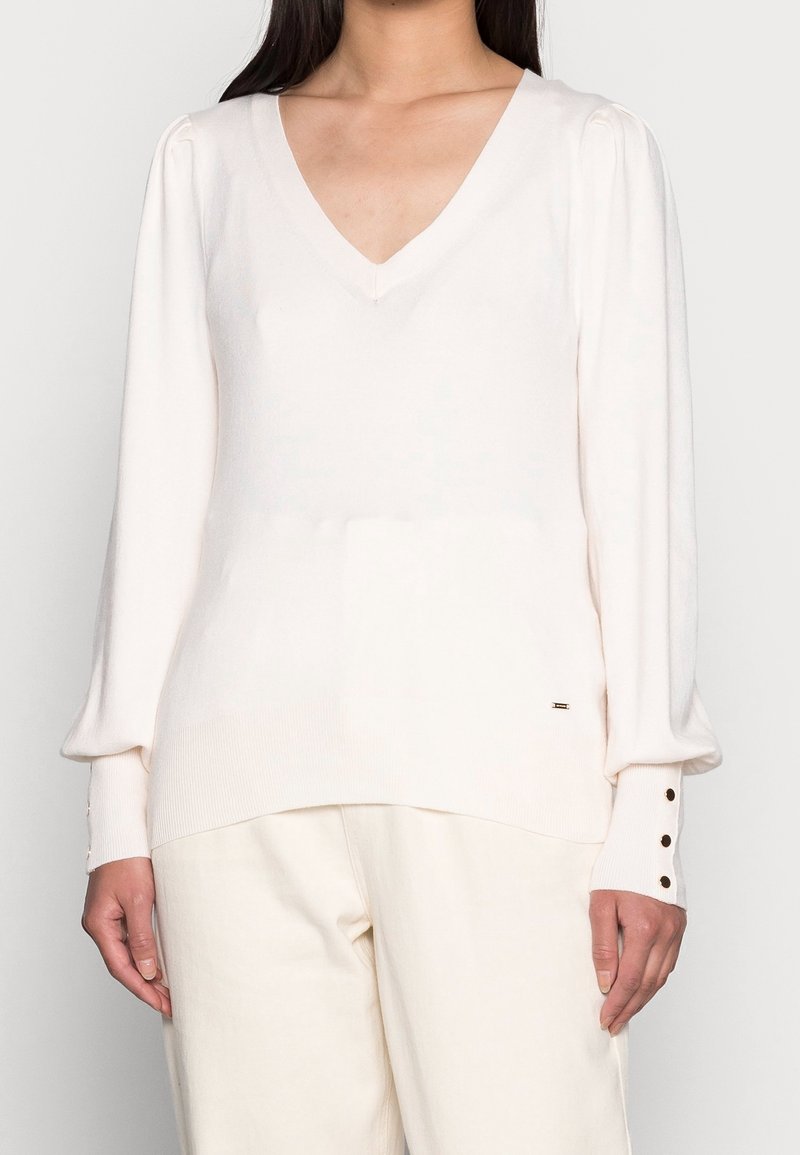 Pull à col en V en maille crème douce avec manches bouffantes. Comprend un ourlet côtelé et des poignets avec des accents de boutons. Associé à un pantalon de couleur claire.