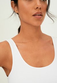 Hvid scoop-neck tanktop med en glat bomuldsstruktur, der har brede stropper og en tætsiddende pasform. Hudfarve synlig i det omkringliggende område.