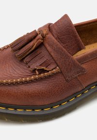 Mocassin en cuir marron avec des pampilles, des coutures détaillées et une semelle noire épaisse avec des accents de couture jaunes.