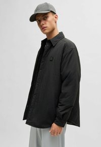 Black button-up shirt with a smooth texture, featuring a small logo patch on the chest. Worn over a black top, paired with gray pants and a cap.