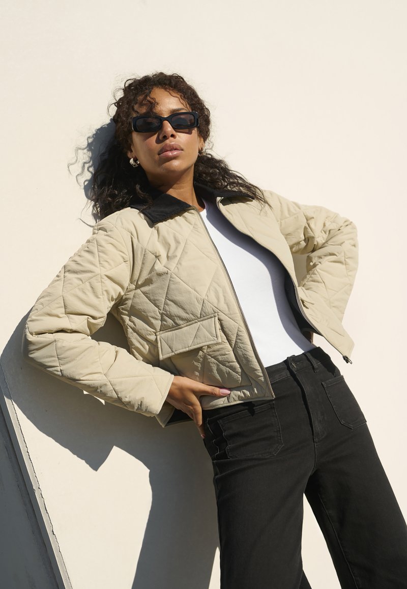 Giubbotto imbottito beige con colletto nero, dotato di tasche frontali. Top bianco sotto e jeans neri a vita alta. Tessuto liscio e texturizzato.