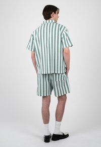 Jeune homme debout, de dos, portant une chemise à manches courtes rayée verticale verte et blanche ainsi qu’un short assorti, avec des chaussures noires et des chaussettes blanches.