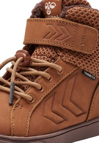 Zapato marrón de caña alta con parte superior texturizada, acentos de cuero, sistema de cordones prominente, correa de velcro y detalle del logo en la lengüeta.