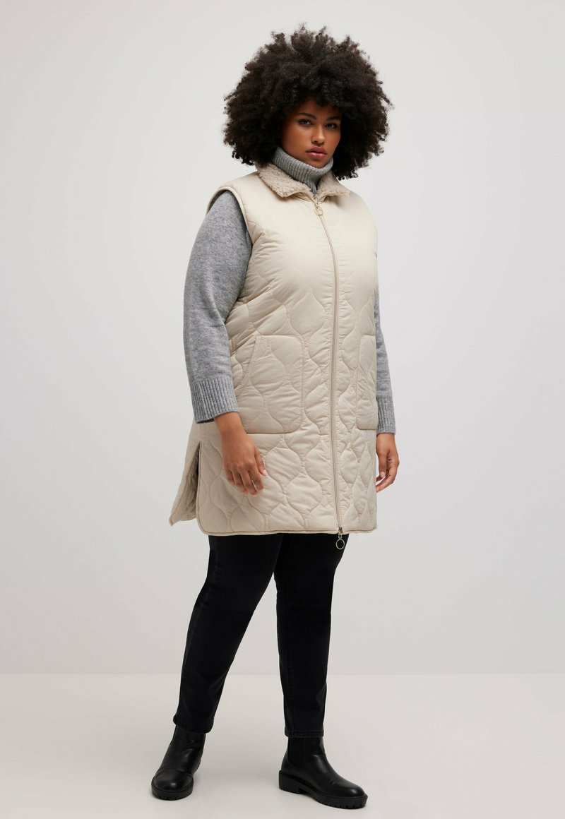 Beige gesteppte ärmellose Weste mit hohem Kragen und Reißverschluss vorne, kombiniert mit einem grauen Rollkragenpullover und schwarzen Hosen, sowie Stiefeletten.