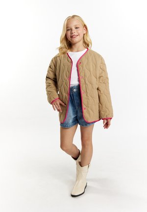 Beige quiltet jakke med rosa detaljer og knapper, båret over en hvid skjorte og denimshorts. Modellen har hvide cowboy støvler på.