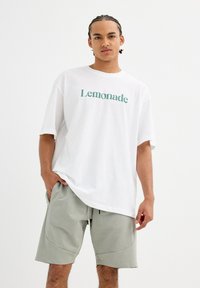 Camiseta blanca con texto verde "Lemonade", combinado con pantalones cortos gris claro de corte relajado y con bolsillos laterales. Textura de tela suave.