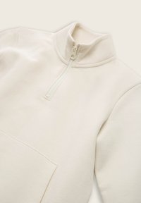 Sudadera con cremallera de color beige claro y cuello alto, con un bolsillo frontal y tela suave y texturizada para mayor comodidad.