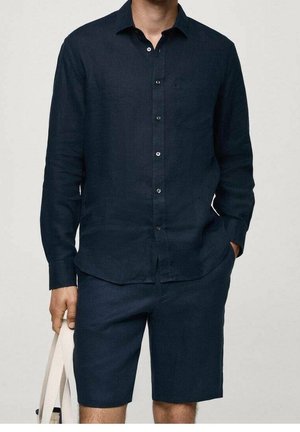 Homme portant une chemise à manches longues bleu marine foncé boutonnée et un short assorti jusqu'aux genoux, tenant un sac fourre-tout crème de la main gauche.