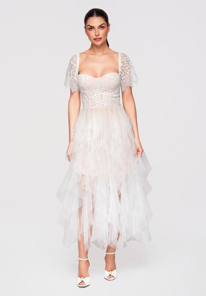 Vrouw draagt een witte kanten korsetjurk met een gelaagde tule rok en korte kanten mouwen, gecombineerd met witte hakken met enkelriempje.