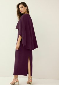 Paarse cape-stijl jurk met een hoge halslijn, vloeiende mouwen, een split aan de zijkant en een gladde textuur. Afgewerkt met metallic sandalen met banden.