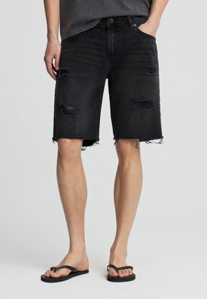 Uomo con pantaloncini di jeans neri consumati con orli sfrangiati e infradito nere, in piedi con una mano in tasca su uno sfondo neutro.