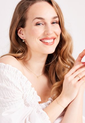 Goldschmuck mit einer zarten Kettenkette, die einen kleinen Anhänger trägt, passenden Ohrringen und Ringen, präsentiert neben einer rüschigen weißen Bluse.