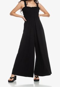 Kvinna som bär en svart byxdress med breda ben och smockad byst, i kombination med svarta plattformssandaler.