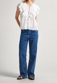 Hvid snøred blouse med volangærmer og bølget kant, parret med bredbenede blå denim jeans og sorte flade sandaler.