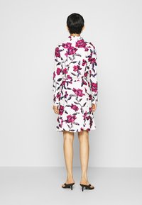 Donna in piedi di spalle, indossando un abito bianco fino al ginocchio con motivo floreale rosa e viola, sandali neri con tacco su sfondo neutro.