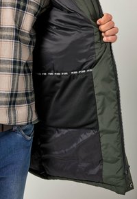 Personne portant une chemise à carreaux et un jean, tenant une veste verte ouverte, montrant une doublure intérieure noire et une poche en maille avec le logo JP1880.