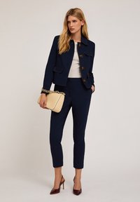 Navyblauwe op maat gemaakte blazer met gouden knopen, witte mesh top, bijpassende op maat gemaakte broek en een beige handtas met een kettingriem.