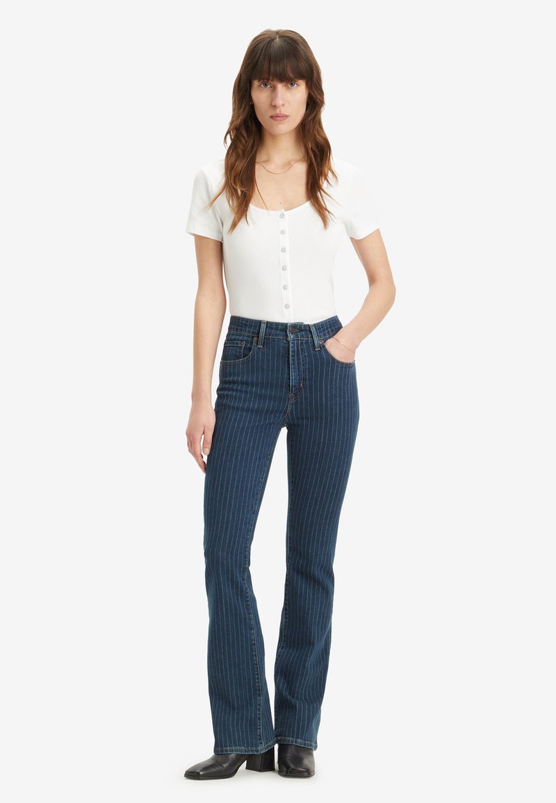 Top blanc à côtes avec des manches courtes et des détails de boutons ; jeans évasés bleu marine à rayures ; bottines noires. Le modèle se tient avec les mains dans les poches.