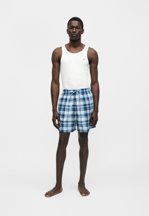 Homme debout pieds nus portant un débardeur blanc sans manches et un short à carreaux bleu et blanc sur un fond clair uni.